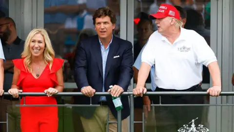Icon Sportswire via Getty Images Marjorie Taylor Greene, smiling in a red dress, Tucker Carlson in a suit coat and khaki pants, and Donald Trump in a white gold shirt and a red MAGA baseball cap stand side by side leaning on a railing