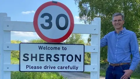 A man in a blue shirt, Wiltshire Cllr Martin Smith, stands beside a 30mph speed sign placed above a 'Welcome to Sherston village' sign. The road is rural with greenery around it.