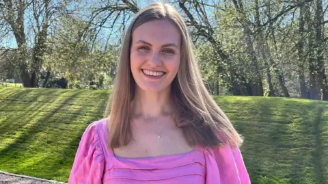 Anna Barker is pictured in a pink blouse. She has long blond hair stood and is stood in front of a lawn