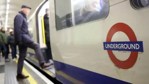 A close-up of a London Underground logo on the side of a train. In the background a man is stepping on to the train.
