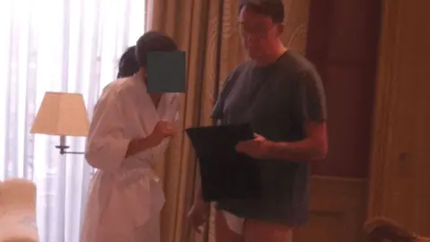 US Department of Justice Mandelson is seen standing next to a woman in a white robe. Her face is redacted. He is wearing a blue T-shirt, and white underwear. He is holding what appears to be an i-Pad.