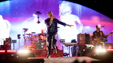 Coldplay on stage at Glastonbury in 2024. Chris Martin is in the front holding a microphone and jumping up. There are purple lights in the background.