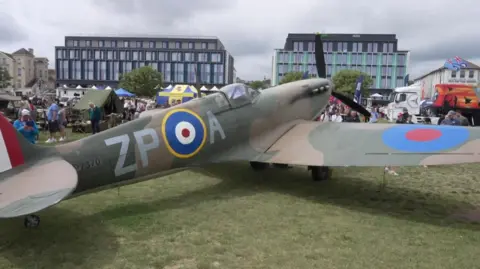A replica Spitfire plane on display on Paignton Green during the second day of the English Rivera Airshow. People are walking around the site which has a number of display tents set up.