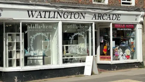 A large shopfront with a white strip above reading Watlington Arcade. Two of the three large windows have stilla hem Swedish Home Style printed on them. The writing on the third window reads Ickle Bitz Toys And Collectibles .