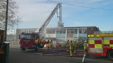 Martin Giles/BBC Two fire engines are outside the derelict building, one has an aerial ladder platform. There are four firefighters in protective clothing walking past one of the fire engines towards the building.