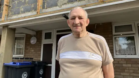 Emily Coady-Stemp/BBC Mike Paternoster standing outside a building with exposed breeze blocks shown on the wall above him. He is wearing a beige Adidas t-shirt.