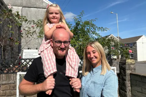 Three people stood in a garden looking at a camera. The man is bald with a beard and glasses, and a black t-shirt. He has a girl with blonde hair on his shoulders. She wears a pink and white top and bottoms. A woman next to him with blonde hair smiles and has a denim shirt on.