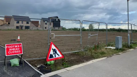 JOANNA TAYLOR/BBC A plot of bare land surrounded by silver metal fencing. In the foreground is a "men at work" road sign and another reading "footway ahead closed". There are houses along one edge of the plot of land. 
