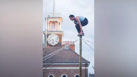 Adam Parkin A man wearing jeans and a vest is balancing on top of a tall wooden pole with ropes coming off it. He has short brown hair and is looking down towards the floor. There is a red-brick clocktower in the background which features a tall white tower.