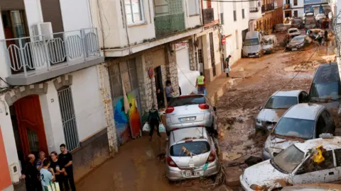 Reuters People gather in a mud-covered street with damaged cars piled on top of each other in Paiporta on 31 October 