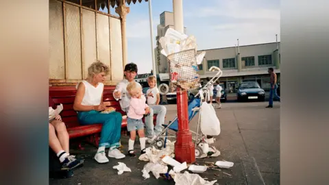 Martin Parr/Magnum Photos A 1980s photo of a family eating fish and chips, with piles of paper rubbish in the foreground