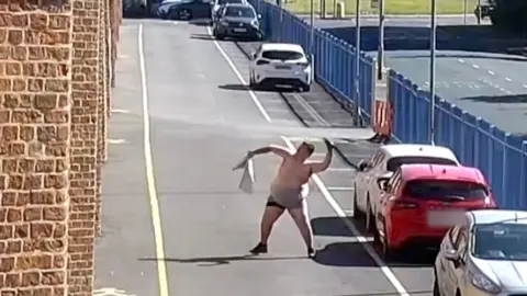 A bare-chested man wearing shorts stands in a street and leans back as he prepares to throw an object into the air. A large blue fence and a tall prison wall are visible at either side of the road along with a row of parked cars.