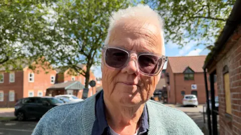 Peter Farnsworth is wearing a light blue jumper with a navy shirt underneath. He is wearing sunglasses with clear frames.