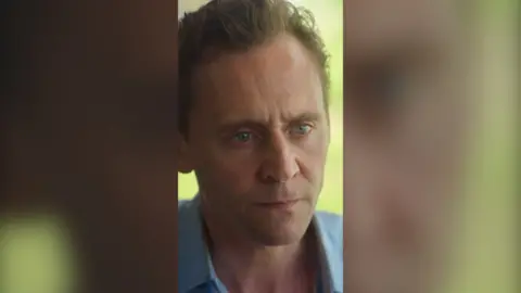Tom Hiddleston's character Jonathan Pine looks angry. He has short blond hair and wears a blue shirt