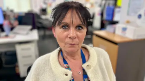 NIKKI FOX/BBC Lucy Kruyer looks at the camera. Her head and shoulders are visible and she is wearing a fluffy cream cardigan. She has a blue company lanyard around her neck. She has deep blue eyes and very dark brown hair with a fringe. Behind her is a blurred background with office furniture, including drawers and desks and a pinboard.
