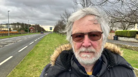 BBC/RICHARD EDWARDS Fairburn parish councillor Derrick Robinson is looking straight at the camera. He has a beard, spectacles and is wearing a blue parka jacket with a fur-lined hood.