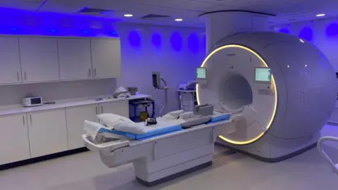 A hi-tech MRI scanner dominates a room in a NHS testing centre. The large machine is in two parts, with a platform, with mattress and pillow, on which the patient is positioned, and a large, cylindrical machine that houses the scanning technology.