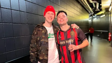 CJM Photography Two men stand in a black corridor. The man on the left wears a camo jacket with  a white T-shirt underneath. He also wears a red woollen hat. The man on the right has dark hair and wears a red and black horizontally striped football top. In his left hand he holds a glass.