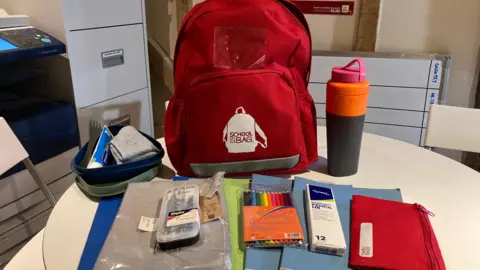 A red backpack sitting on a white round table. In front of it there are a selection of books and folders, a pencil case, washbag, school supplies and a water bottle.