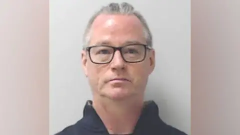 Police Scotland Man with short greying hair, wearing dark-rimmed glasses, looking at camera, wearing a dark top, in a police mugshot.