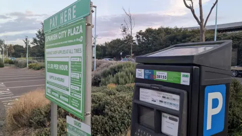 BBC/Leigh Jones A black solar paneled pay and display machine stands in the foreground of a car park surrounded by tidily kept shrubbery. Next to it stands a green and white sign entitled 'pay here' with instructions and terms and conditions for parking on the site. 