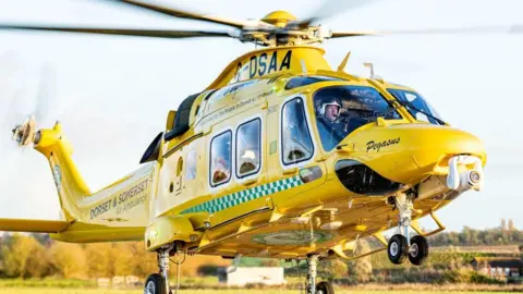 A Dorset and Somerset Air Ambulance, painted bright yellow, rises just above the ground. The face of the pilot is just visible through the windscreen.