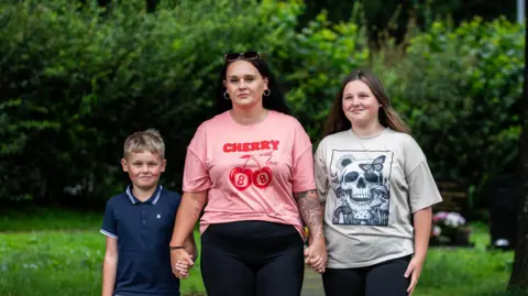 SWNS Photograph of Hazel Jones (centre) pictured with her daughter Amelia, 13, (right) and her son (left). Hazel Jones wears a pink t-shirt with red writing on the front which reads "cherry". Her daughter wears a grey t-shirt with a navy skull design, while her son wears a navy polo shirt and has short blonde hair. 