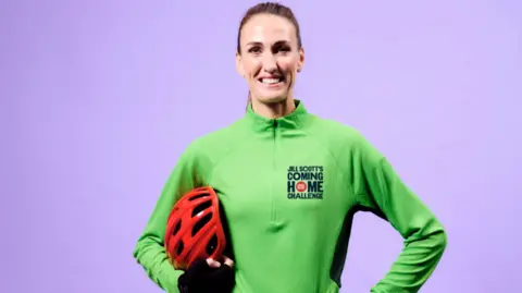 Comic Relief/PA Jill Scott is standing in front of a purple background. She is smiling widely as she holds a red helmet under her arm. She is wearing a green quarter-zip top with Jill Scott's Coming Home Challenge written on it. Her hair is slicked back in a pony tail.