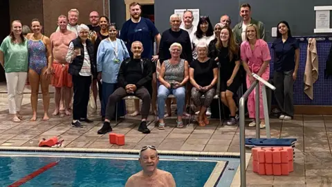 Ken is in the water in the indoor swimming pool. He has swimming goggles on his head. Behind him are friends and family to cheer him on along with Kay.