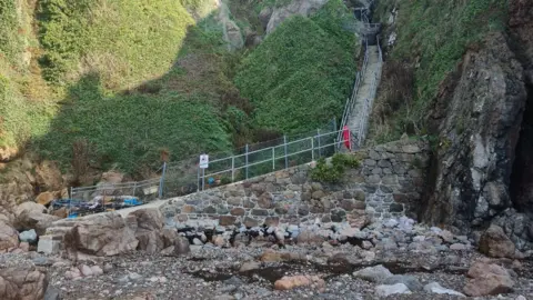 The bottom of the Petit Port steps leading on to a stony beach. There are metal handrails, fencing with a sign showing a hazard warning of falling rocks and a life buoy. The steps lead sharply up a stiff grassy cliff face. 