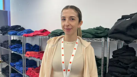 Paula wearing a beige hoodie and lanyard in front of a row of different coloured uniform.