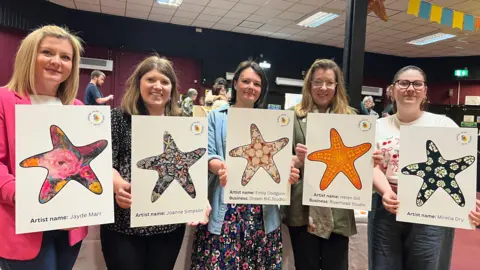 Five women are smiling into the camera and holding up white posters of starfish designs. It includes pink and yellow roses, shells on the sea and an orange flowery design.