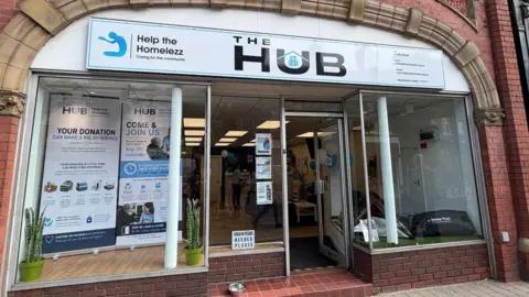 A photo of the front of the centre, which has a glass front with the logo above on a panel. The door is open and there are plants in the windows and information about how people can donate.