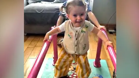 A young girl with short blond hair, with small pigtails, has her hands resting on two parallel metal pink bars, which are attached at the bottom to a walking mat. She is walking and has a beaming smile. She is wearing yellow striped trouser bottoms and has a light yellow vest. 