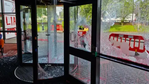 Aftermath of an attack by protestors on a BBC building. A revolving door and panels with shattered glass, some of which has also fallen onto the floor, and red spray paint. Outside, barriers have been erected.