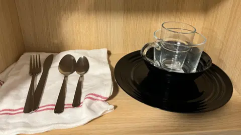 A wooden shelf with a white and red striped tea towel, a fork, knife, spoon and teaspoon, a water glass, a mug, a black bowl and a black plate.