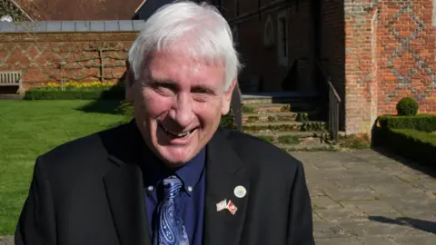 Bobby Dyer has grey hair and appears to be laughing. He is wearing a black suit jacket, navy shirt and navy tie. He is in the grounds of Layer Marney Tower.