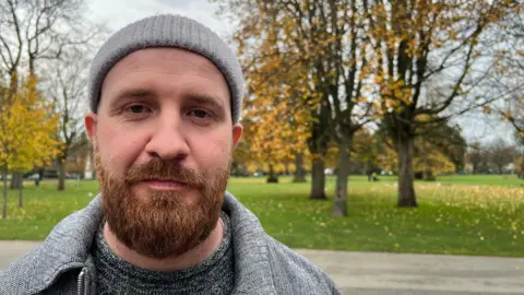 A man with a full brown beard stands in a park in front of a large grassy field and mature trees with autumn leaves. He is wearing a grey coat, grey jumper and grey beanie hat.