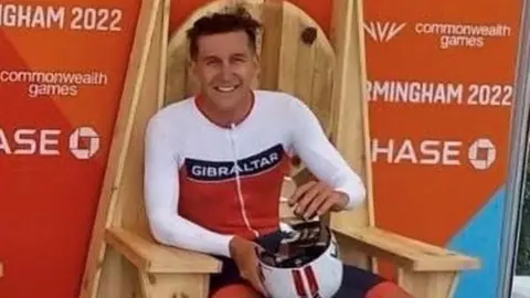 Family handout A smiling man sat down on a wooden chair. He is wearing a white and orange sports top which has 'Gibraltar written on it' and is holding a white cycle helmet. 