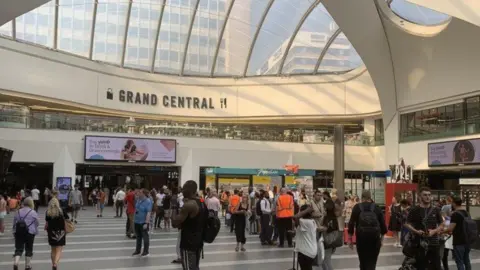 Approximately 30 people spread out across the station concourse on a typical day at New Street. Above them is a clear roof revealing the city-scape beyond. Some of the people face an arrivals and departures board which is out of shot.