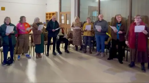 A number of people of different ages are standing in a semi-circle and are holding a piece of paper as they are singing. 