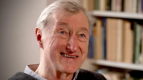 BBC/Adam Walker Julian Barnes smiling, top of checked shirt collar & dark top. Book shelf behind him.