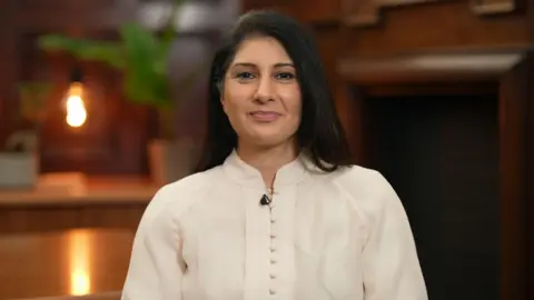 Aliya is wearing a cream, buttoned blouse and has long, black hair. She is smiling at the camera and is sat in a room with oak-lined walls. In the background, blurred, are a desk, lamp and plant.