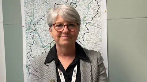 Kate Bradbrook/BBC A woman wearing glasses and a back and white chequered blazer and black open shirt stands in front of a map of Northamptonshire. 