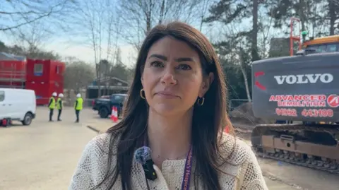 Ben Parker/BBC Laura Jenkins with long dark hair. She is wearing a cream top. In the background are construction vehicles and workers.