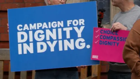 PA Campaigners holding up placards outside the Manx parliament. A blue sign says Campaign for dignity in dying, while a pink one has the words Choice, Compassion, Dignity listed with ticks next to them.