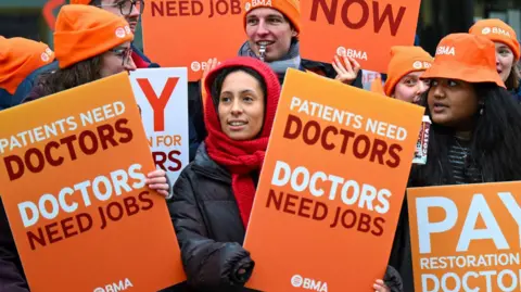 Resident doctors stand together outside wearing winter coats and hats while striking, holding orange placards saying 'doctors need jobs' and 'pay restoration now'