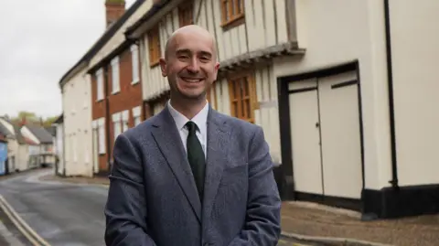 Ben Goldsborough is wearing a grey suit and standing outside a row of homes, one of which appears to be from the tudor-era.