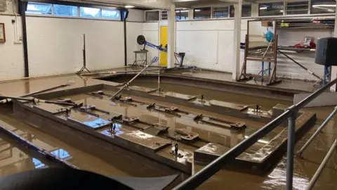 The inside of a room with several rowing machines, which are sitting in dirty brown flood water.