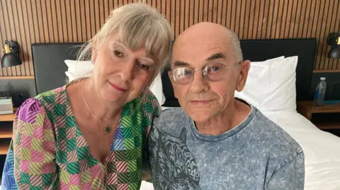 Karen and Colin McGrory are staying in a hotel after not being allowed back into Mercia House. They are sitting in a hotel room. Mrs McGrory is wearing a colourful top and a pendant necklace and has her hair tied back. Mr McGrory is wearing glasses and has a blue T-shirt.